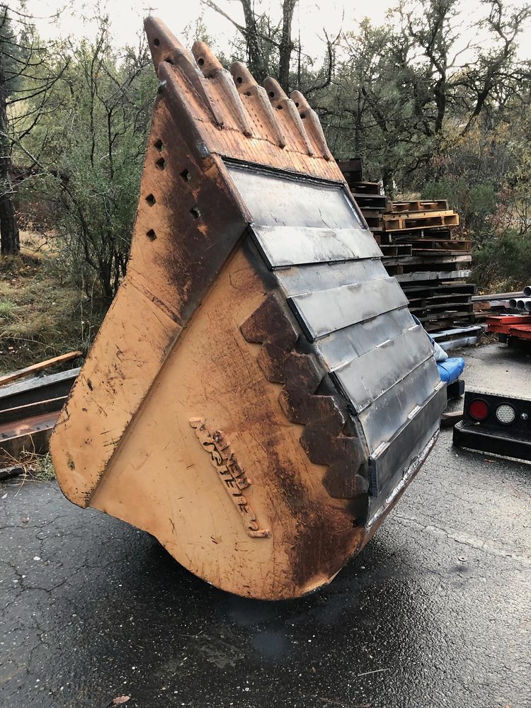 Excavator bucket rebuilt with new wear plates and side cutters, standing on asphalt outside the shop, pines behind